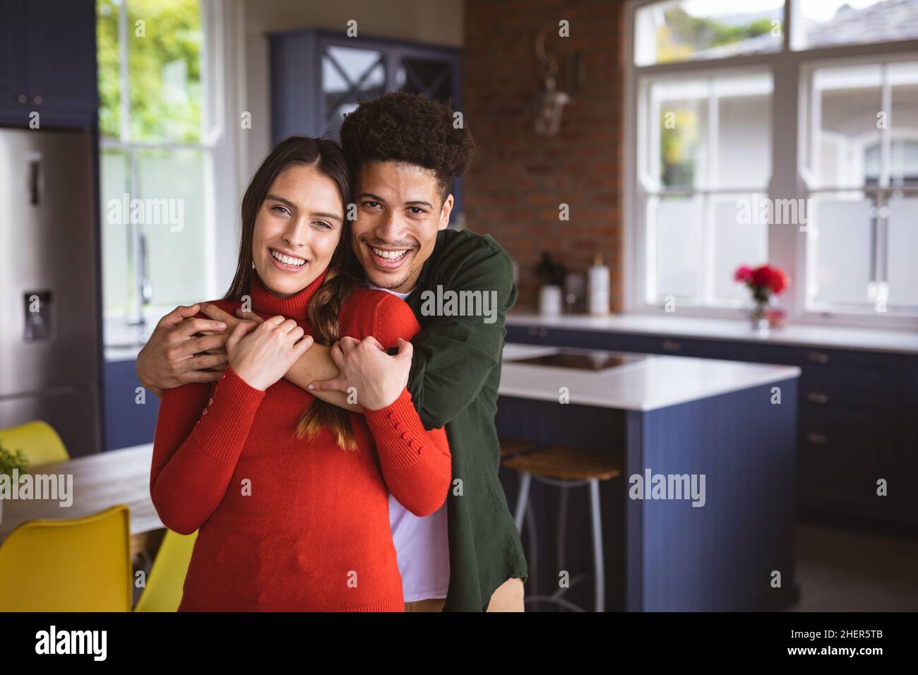 Porträt eines glücklichen jungen Birazialmannes, der die Freundin von hinten umarmt und zu Hause in der Küche steht Stockfoto