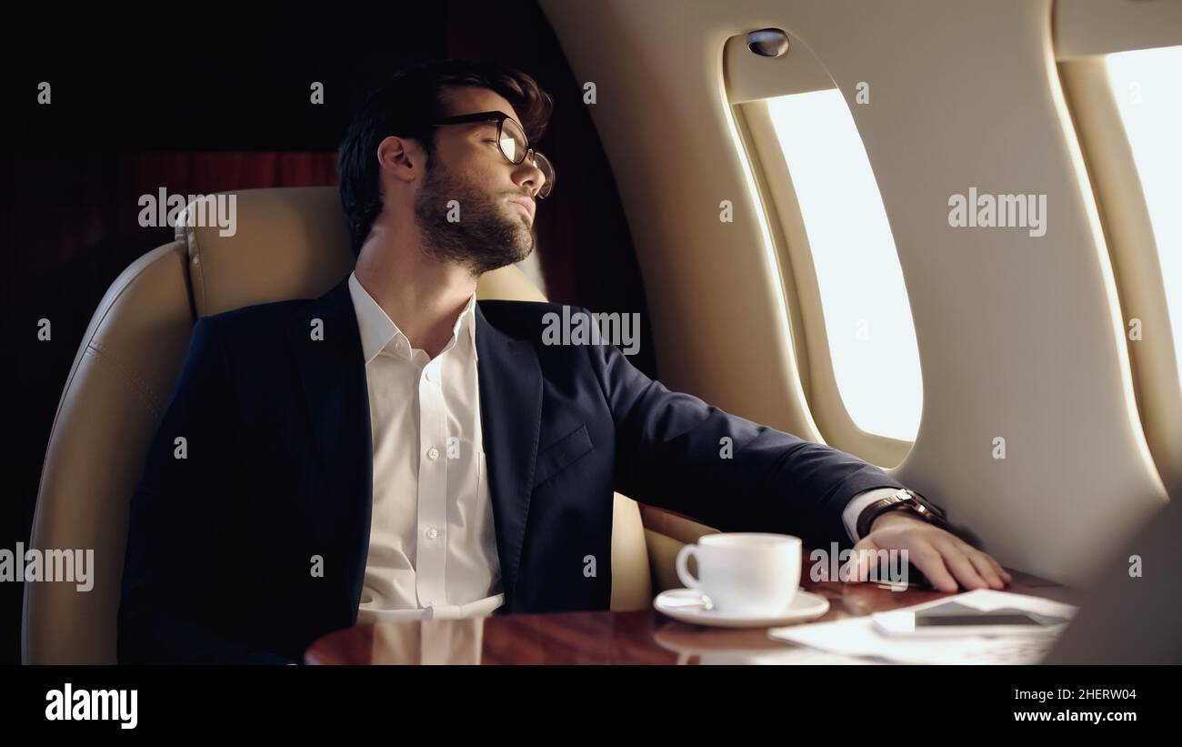 Geschäftsmann, der im Privatjet in der Nähe von Kaffee und Smartphone auf das Fenster blickt Stockfoto