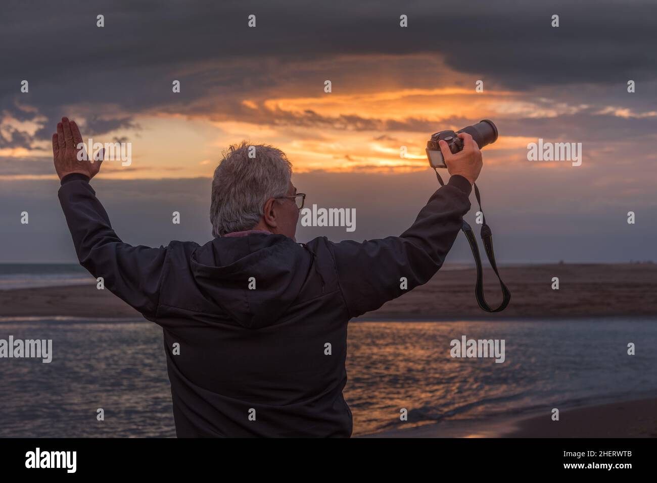 Reifer erwachsener Mann mit erhobenen Armen und Kamera in der Hand, der in den Sonnenuntergang blickt Stockfoto