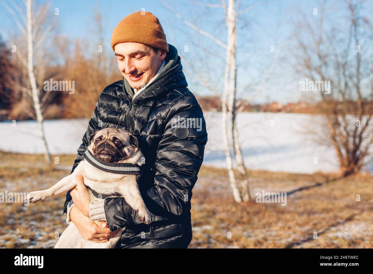 Junger Mann, der im verschneiten Winterpark einen Mops-Hund spaziert. Der Meister spielt mit einem Haustier, das es an den Händen hält. Aktivitäten mit Tieren Stockfoto