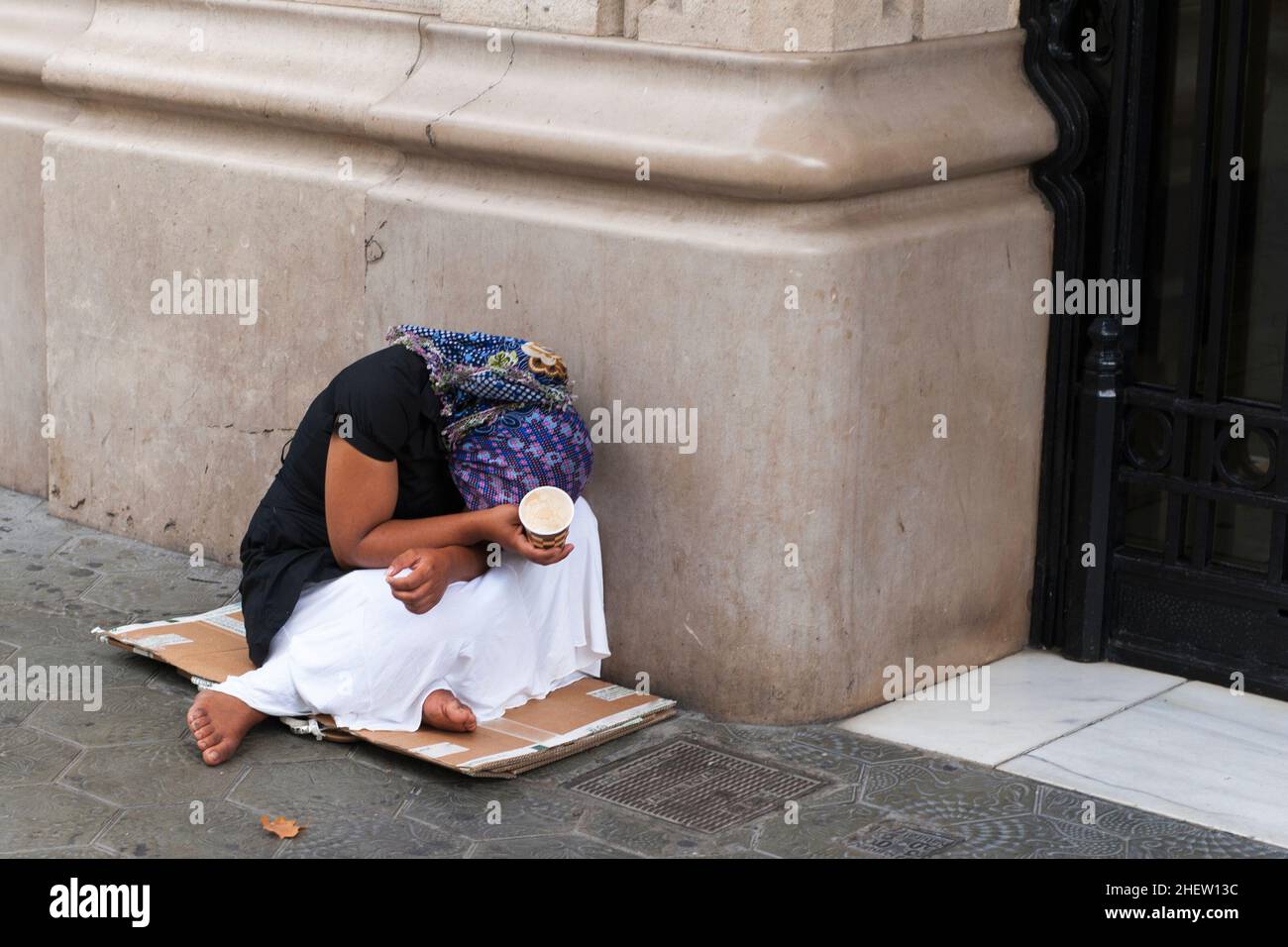 Barfuß verarmte Bettlerin, die sich auf einem alten Stück Pappe vor einem Gebäude mit verborgenem Gesicht und einem Bettelbecher in ihrem Haa kauerte Stockfoto