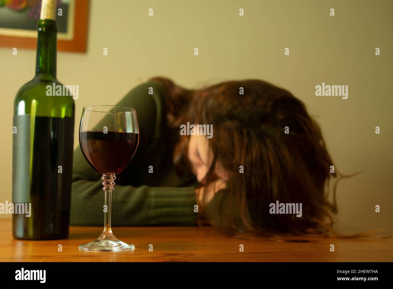 Nahaufnahme einer Frau mit deprimiertem Ausdruck in einer grünen Jacke, die mit ihrem Kopf auf dem Tisch sitzt, begleitet von einer Flasche und einem Glas Rotwein Stockfoto