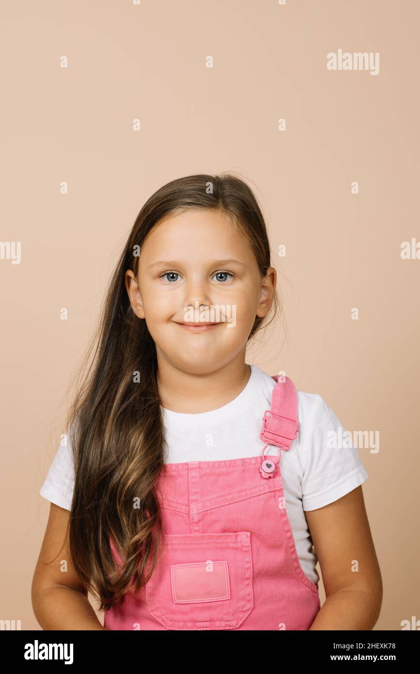 Close up Face Kind mit leuchtend leuchtenden Augen und ruhigen glücklich breiten Lächeln Blick auf die Kamera in leuchtend rosa Jumpsuit und weißem T-Shirt auf beige Stockfoto