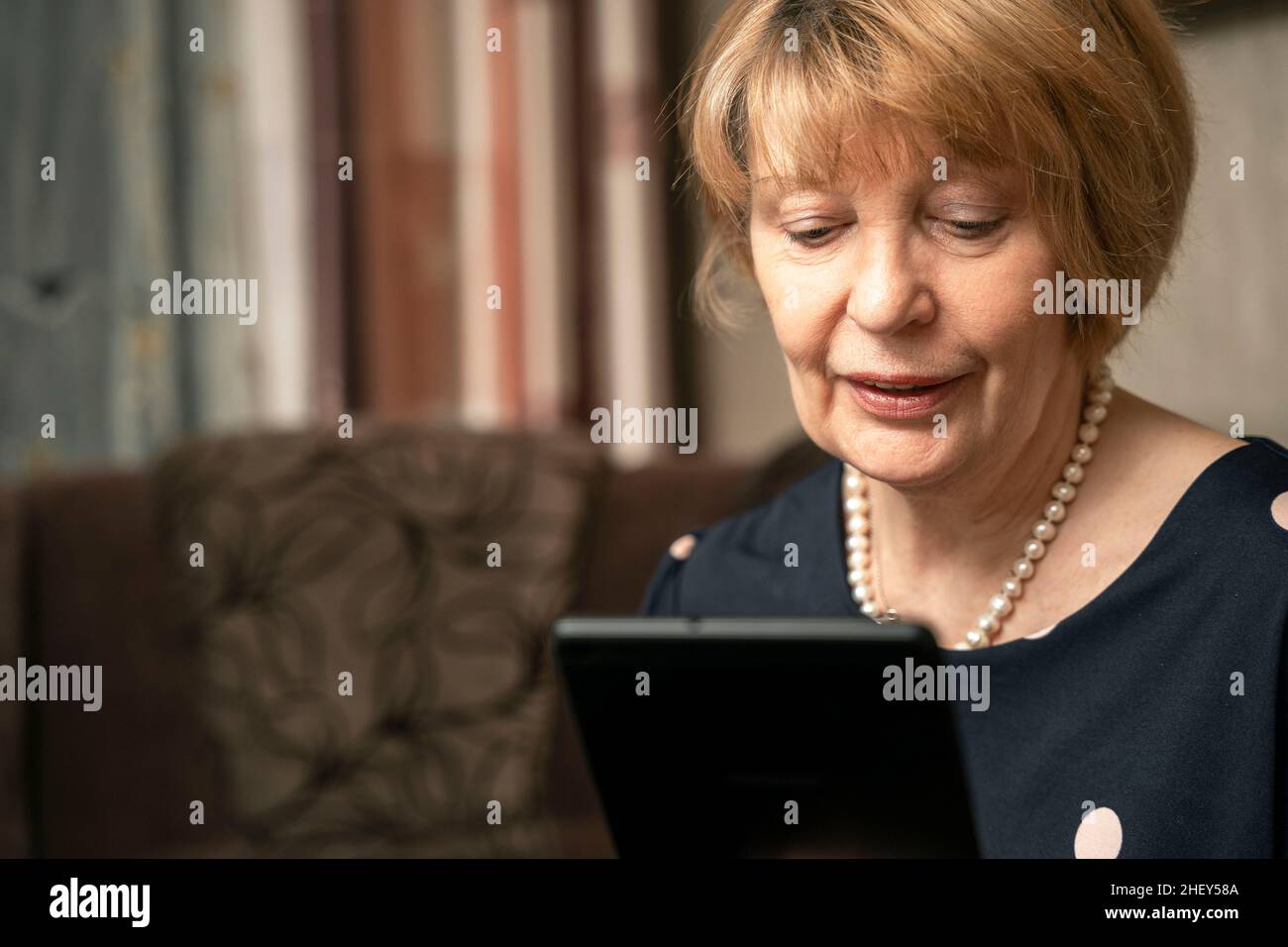 Eine ziemlich ältere Frau benutzt ein Tablet und das Internet, während sie im Wohnzimmer auf einem Sofa sitzt. Süße Heimat, Großmutter kommuniziert mit ihrer Großmutter Stockfoto