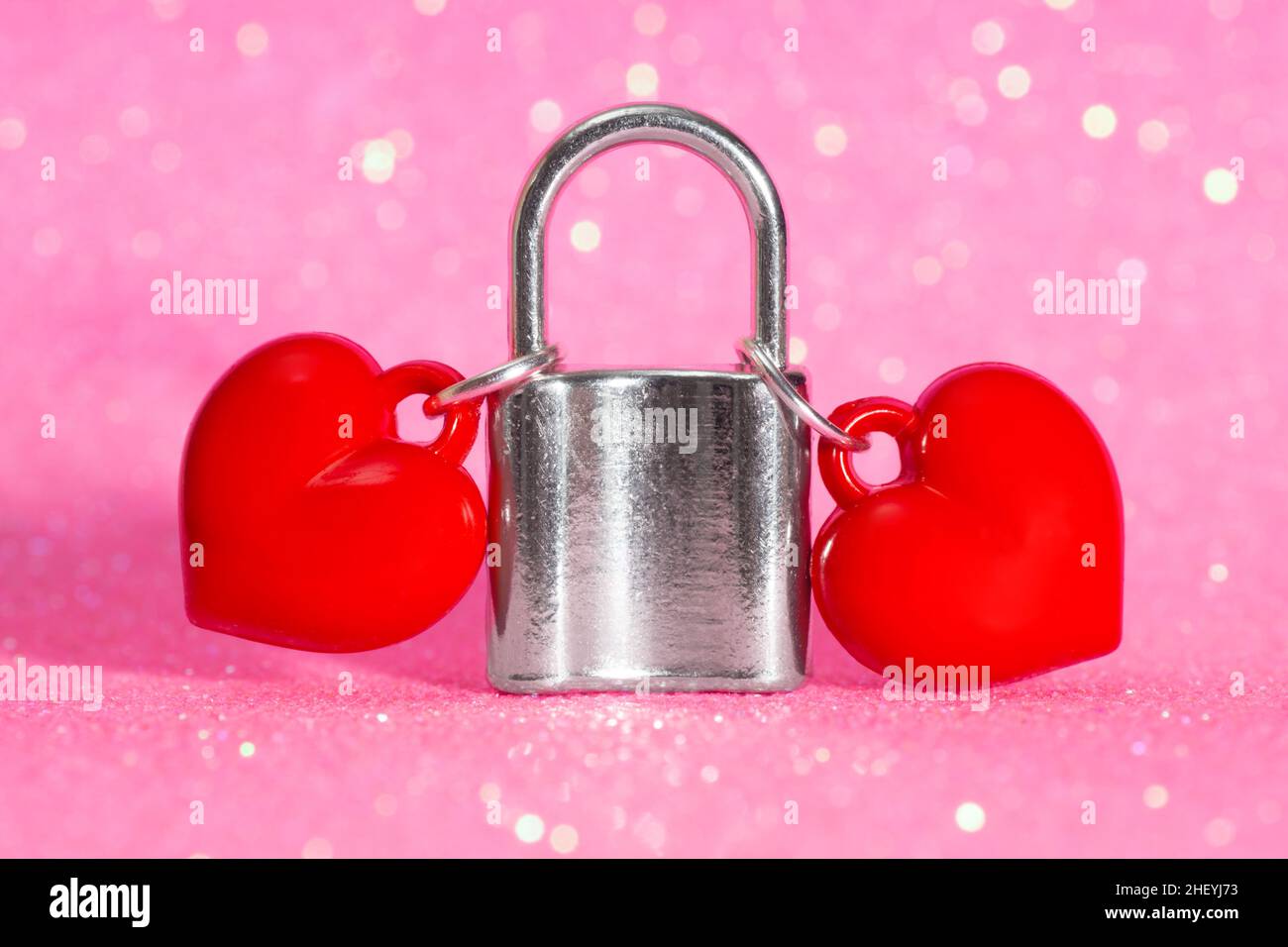 Zwei rote herzförmige Anhänger, die an einem silberfarbenen Vorhängeschloss auf einem glitzernden rosa Hintergrund befestigt sind. Das Konzept der ewigen Liebe zwischen einem Paar Stockfoto