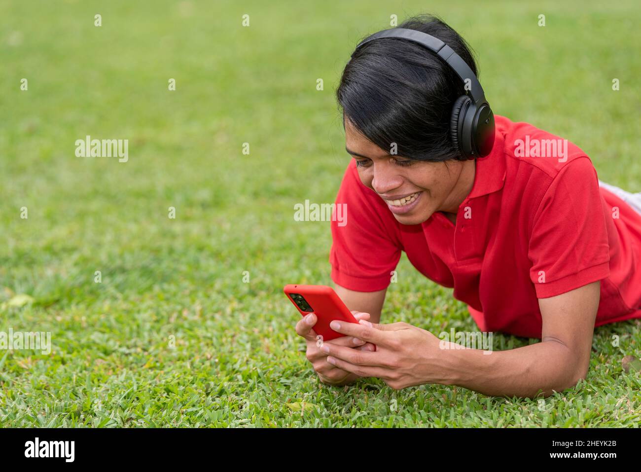 Fröhlich lächelnder junger Latino-Mann, der mit dem Smartphone auf dem Gras liegt. Stockfoto