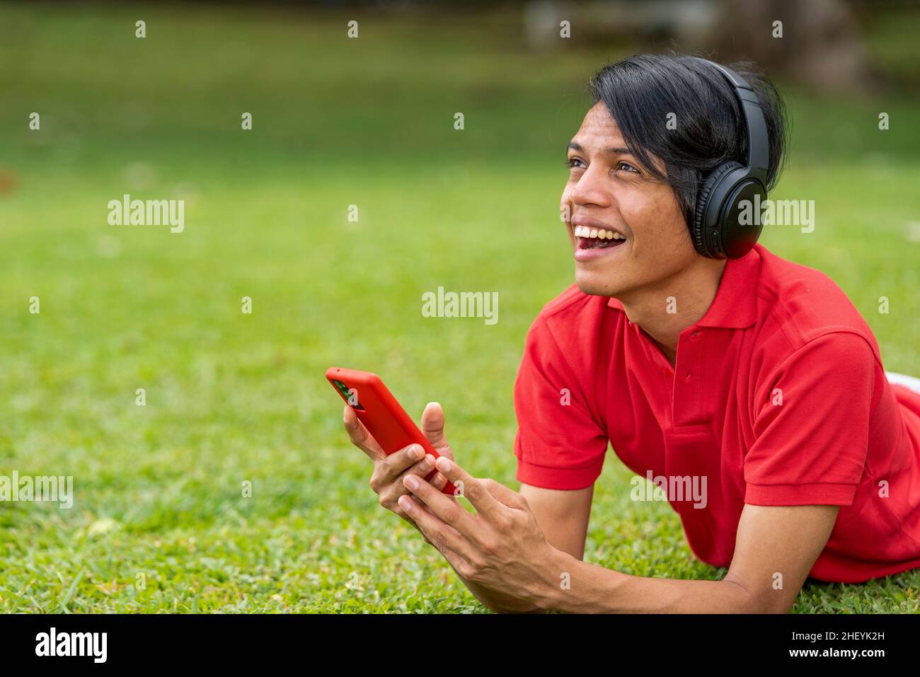 Fröhlich lächelnder junger Latino-Mann, der mit dem Smartphone auf dem Gras liegt. Stockfoto