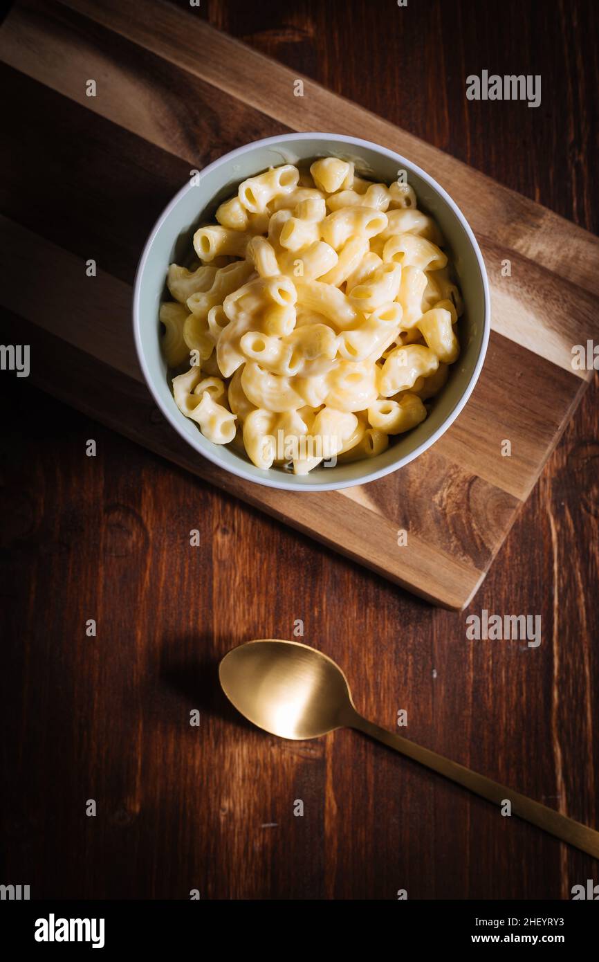 Klassischer mac und Käse mit Basilikum auf der Oberseite und mac und Käse im Ofen im Hintergrund mit Gabeln und rohen Makkaroni auf einem rustikalen Holzhintergrund gebacken Stockfoto