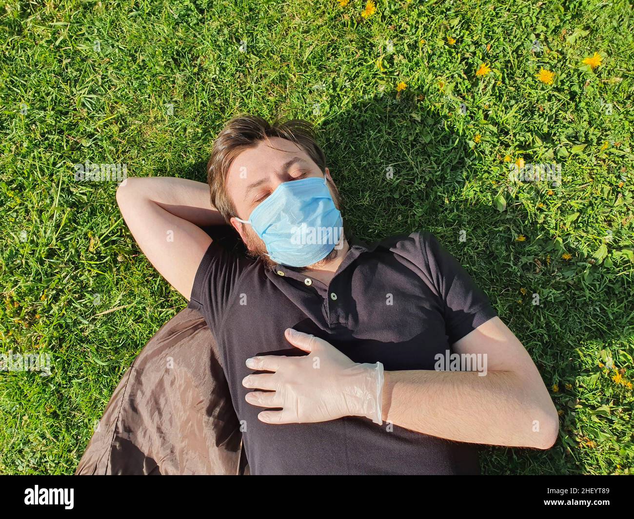 Kaukasischer junger Mann mit medizinischer Gesichtsmaske und medizinischen Handschuhen liegt im Gras im Park. Schutz Ihres Gesundheitskonzepts. Lifestyle Stockfoto