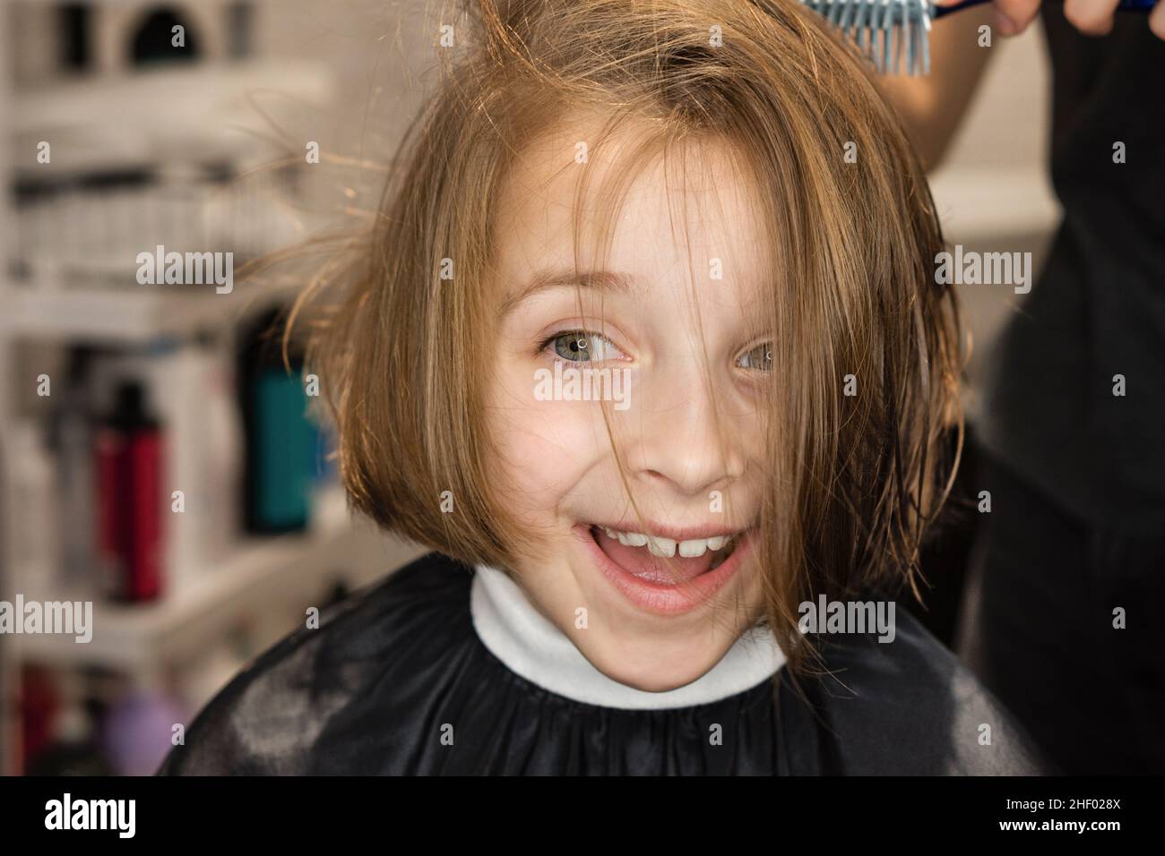 Angenehm überrascht, dass das Kind im Schönheitssalon einen Haarschnitt bekommen hat. Friseur mit Trockner nass Schuss Haar im Friseursalon. Kleines Mädchen mit dunkelblondem bob beim Stylist. Stil und Mode für Kinder. Stockfoto