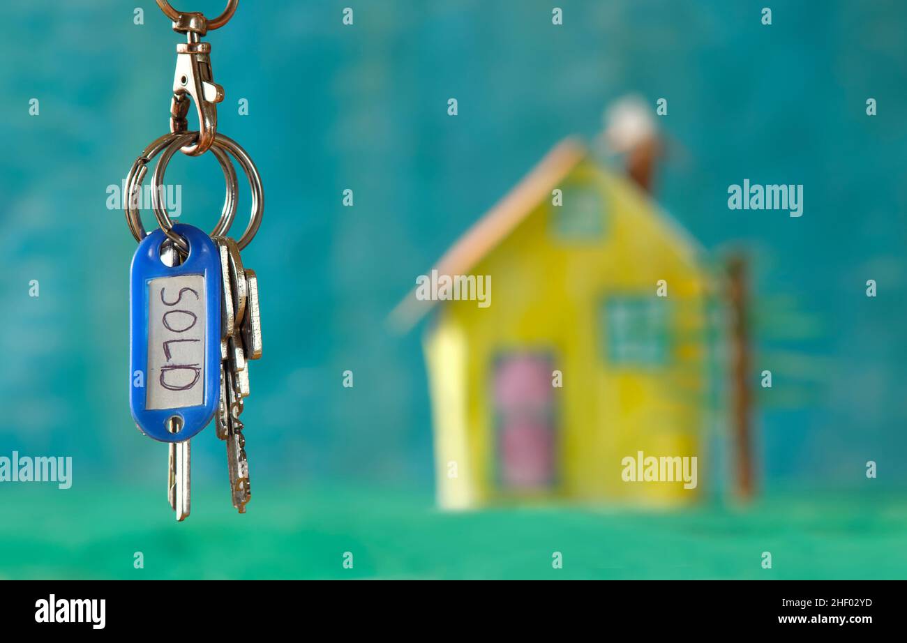 Schlüsselanhänger mit Schlüsselanhänger und verkaufter Markierung, verschwommenes Modellhaus auf der Rückseite, Haus kaufen, Immobilienkonzept. Selektiver Fokus, freier Kopierraum Stockfoto