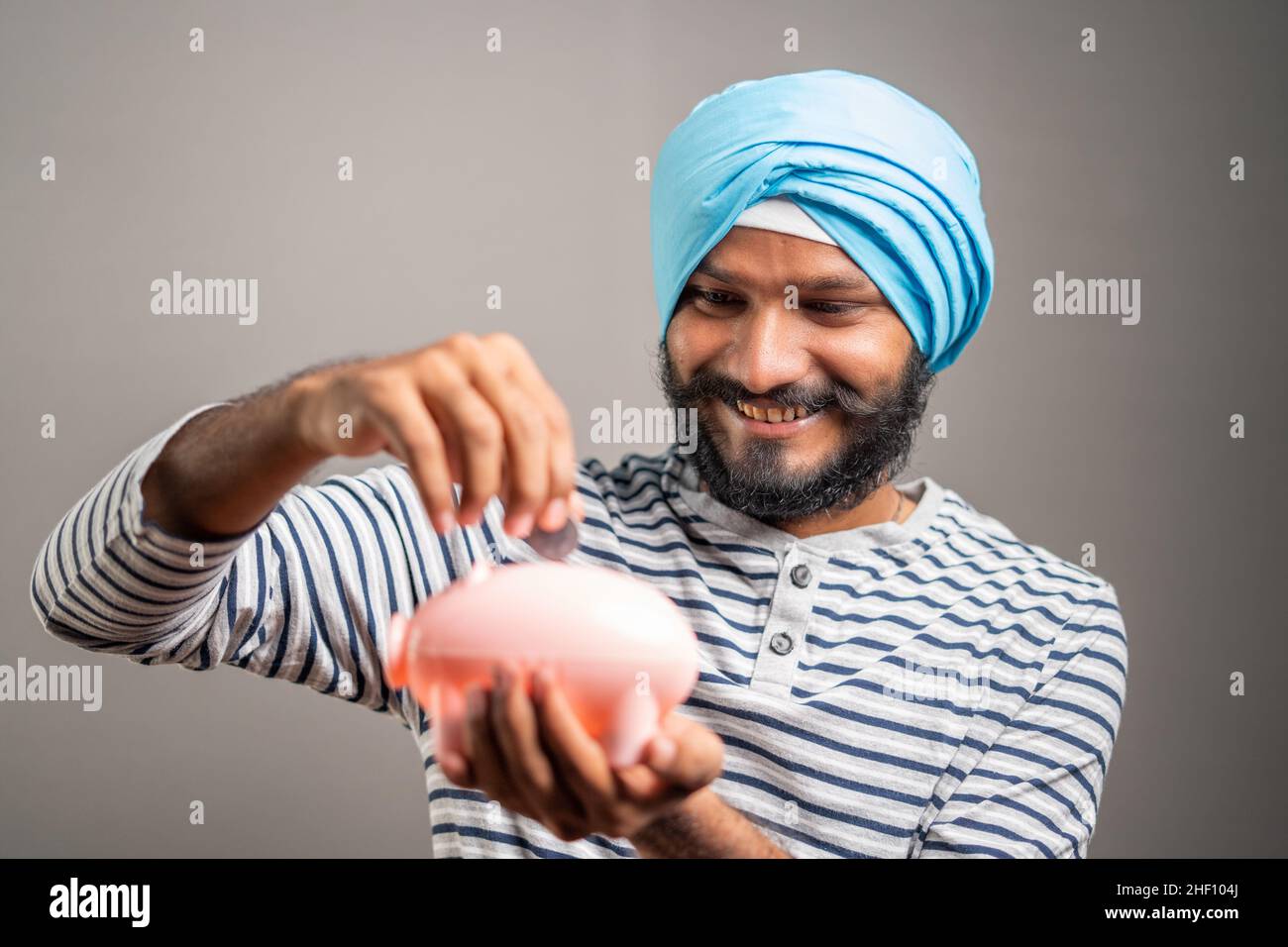 Junger indischer sikh-Mann spart Geld, indem er auf dem Sparschwein auf grauem Hintergrund eine Münze legt - Konzept des Sparens und der zukünftigen Finanzplanung und -Investition. Stockfoto