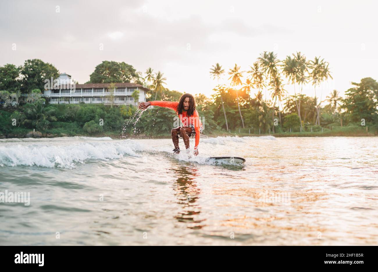 Schwarzer langhaariger Teenager-Junge, der ein langes Surfbrett reitet. Er fing eine Welle in einer Bucht des indischen Ozeans mit magischem Sonnenuntergang Hintergrund. Extreme Wassersportarten und ex Stockfoto