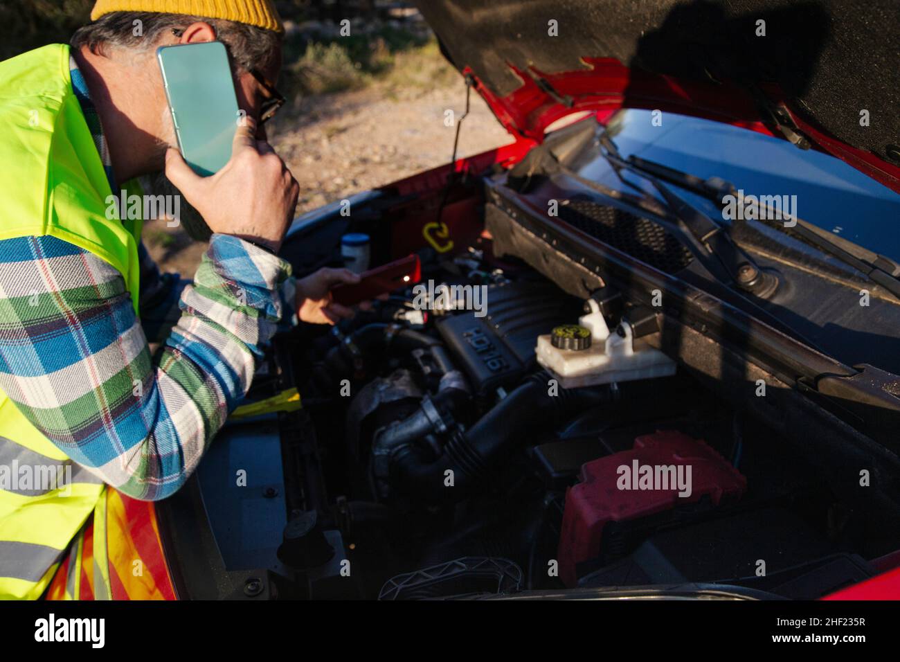 Kaukasischer Mann, der unter der Haube eines kaputten Autos mit einem Mechaniker telefoniert. Stockfoto