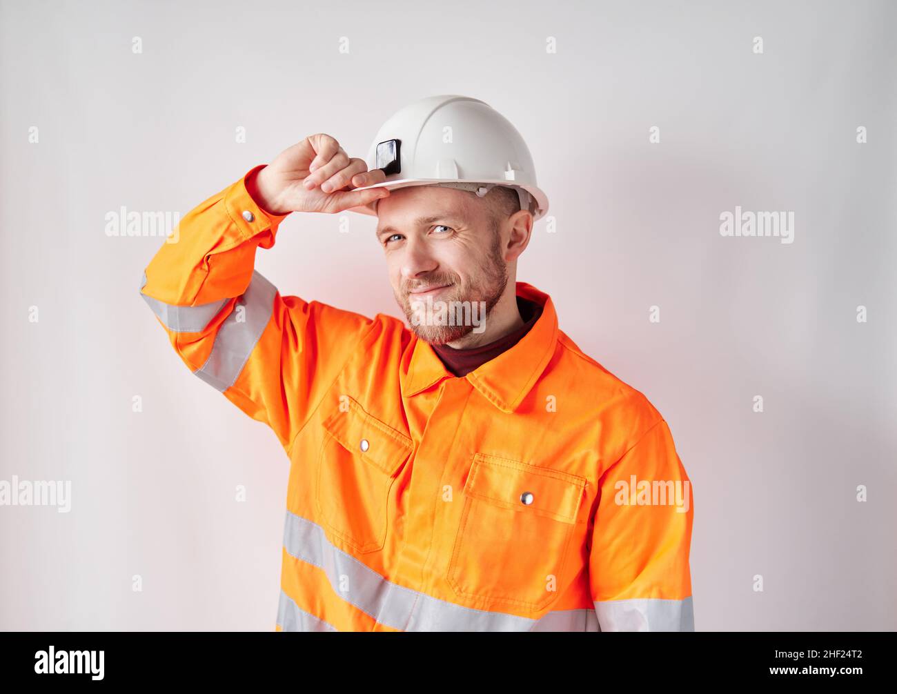 Lächelnd und schauend auf Kamera niedlichen kaukasischen Techniker, Ingenieur, Supervisor oder Bauunternehmer in weißem Helm und orange Jacke Korrektur seines Arbeitshelms. Konzept der Bauindustrie Stockfoto