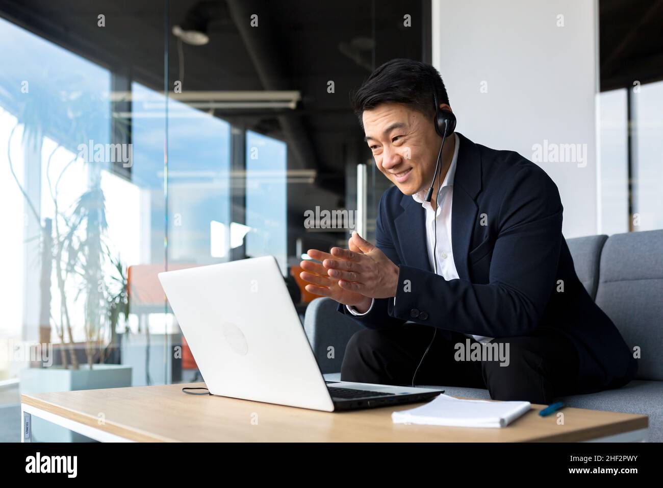 Ein Mitarbeiter im asiatischen Callcenter spricht mit Kunden im Büro, ein asiatischer Geschäftsmann nutzt ein Headset und einen Laptop für einen glücklichen und lächelnden Videoanruf Stockfoto