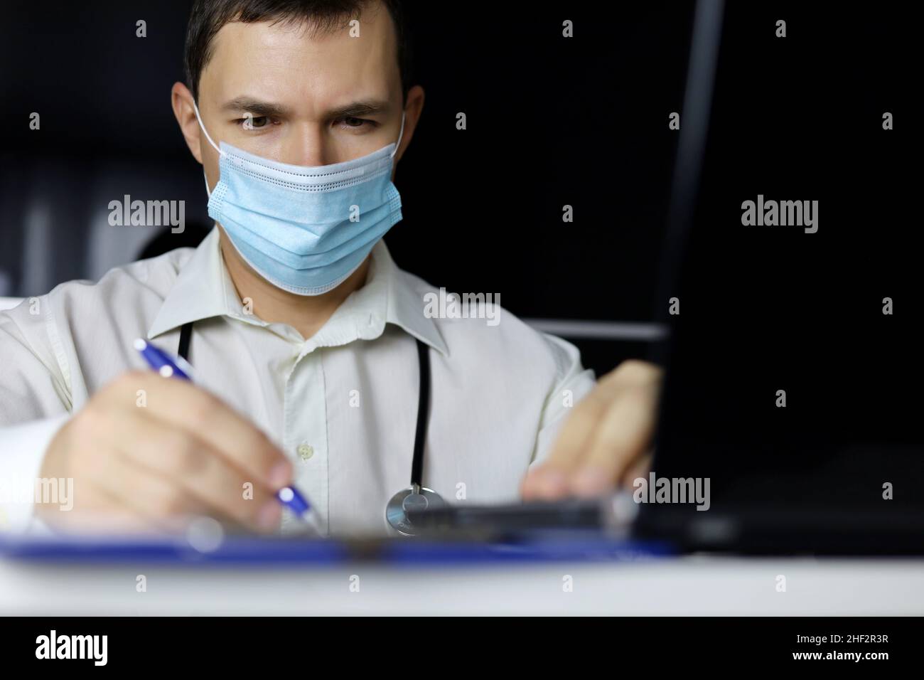 Arzt in Einwegmaske schreibt verschreibungspflichtiges Papier sitzen am Laptop im Büro. Arzt bei der Arbeit, Konzept der medizinischen Untersuchung, Ausfüllen der medizinischen Stockfoto