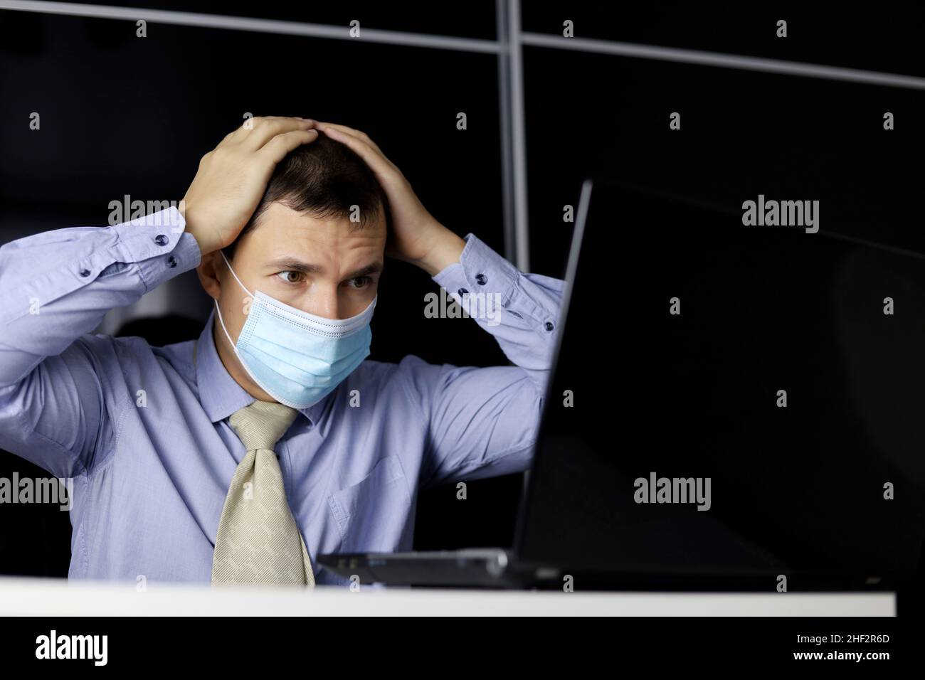 Mann in Gesichtsmaske und Bürokleidung, der aufgeregt auf das Laptop-Display schaut und seinen Kopf mit den Händen hält. Konzept der Lösung einer schwierigen Aufgabe, Fehler bei der Arbeit Stockfoto