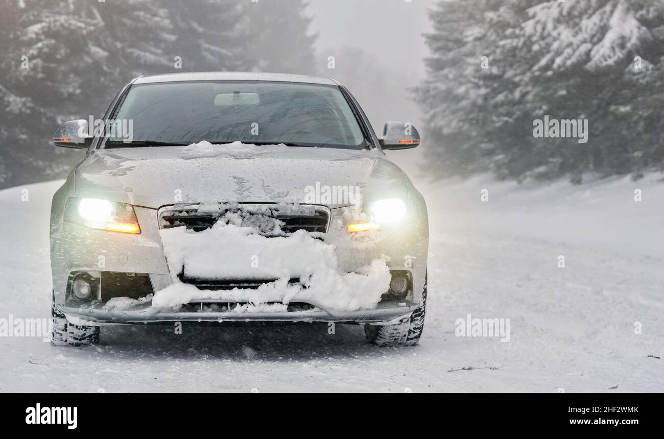 Silbernes Auto auf schneebedeckter Winterstraße geparkt, verwackelte Bäume Hintergrund, Vorderansicht, Scheinwerfer leuchten Stockfoto