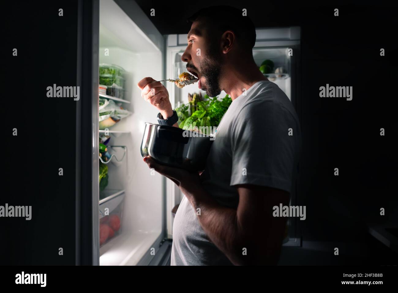 Junger hungriger Mann, der nachts aus dem offenen Kühlschrank Essen isst. Mitternachtsimbiss und Essensabhängigkeit Stockfoto