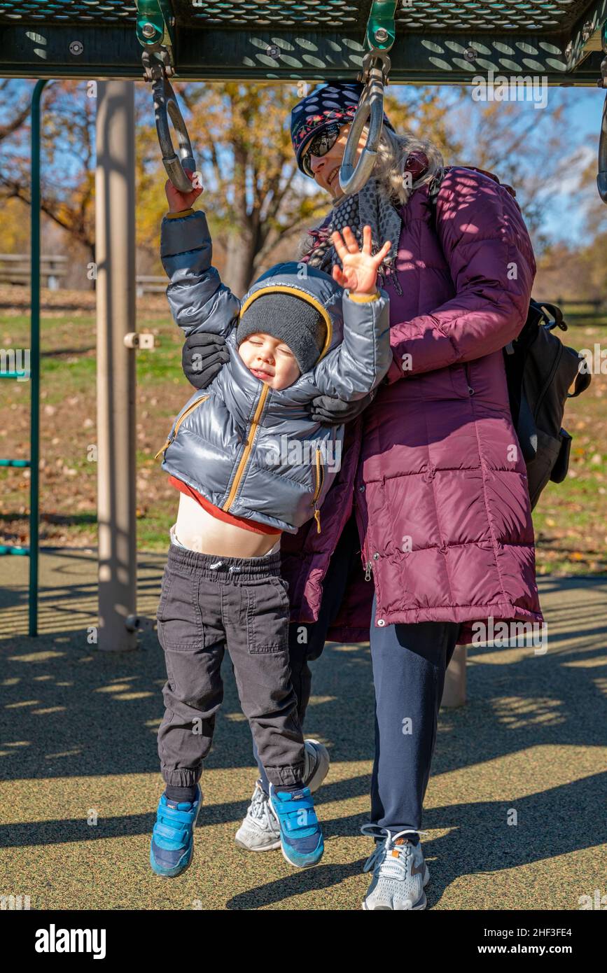 Großmutter hilft einem zweijährigen Jungen, der auf dem Spielplatz im Stadtpark hängt Stockfoto