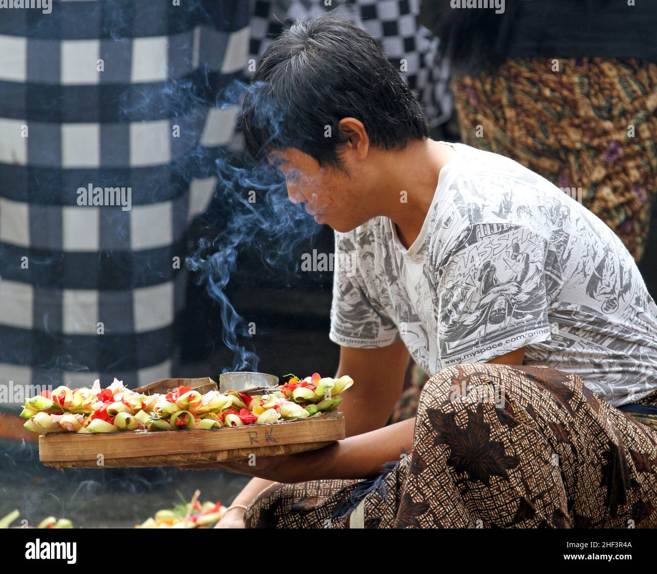 Ein junger balinesischer Mann hockte sich und trug ein Rattantablett mit Canang Sari Hindu-Opfergaben auf dem Sukawati Markt in Bali, Indonesien Stockfoto