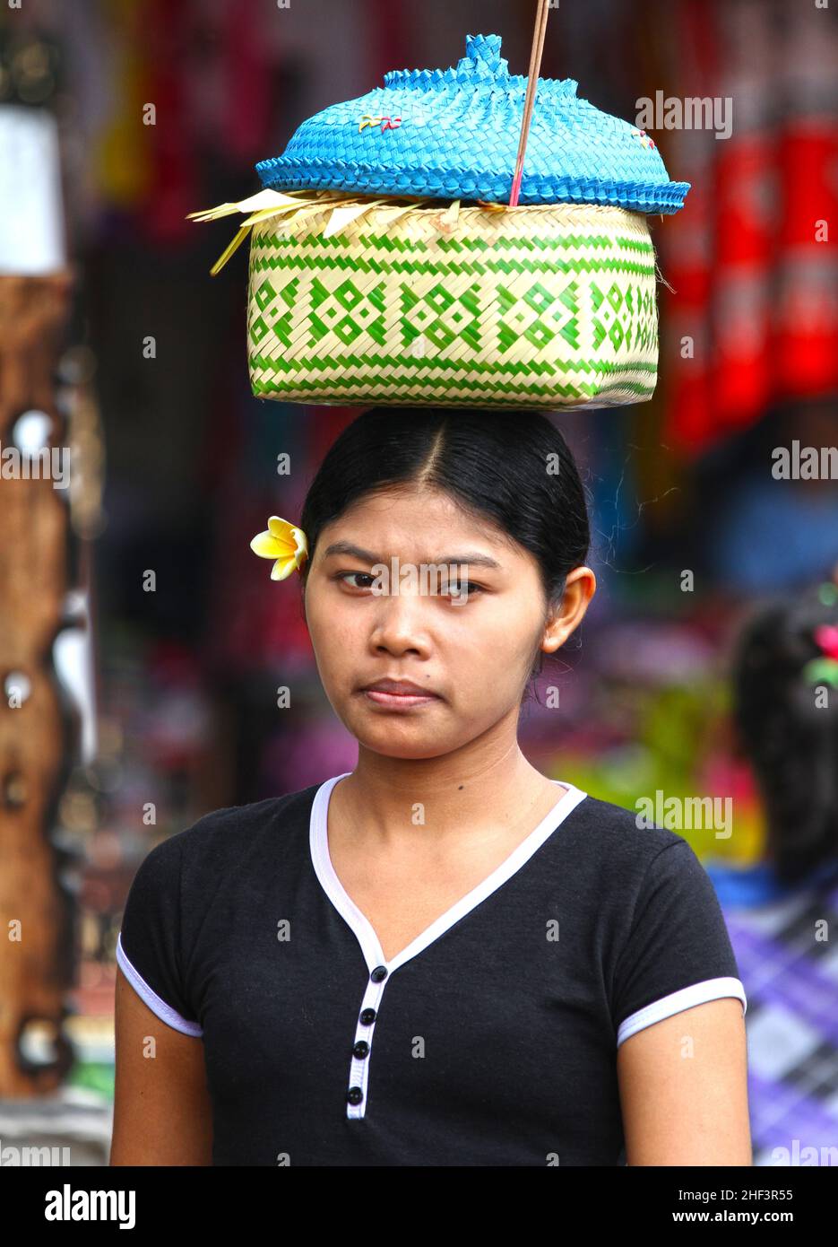 Eine junge Balinesin mit einer Frangipani-Blume im Haar, die einen Korb auf dem Kopf auf dem Sukawati-Markt in Bali, Indonesien trägt Stockfoto