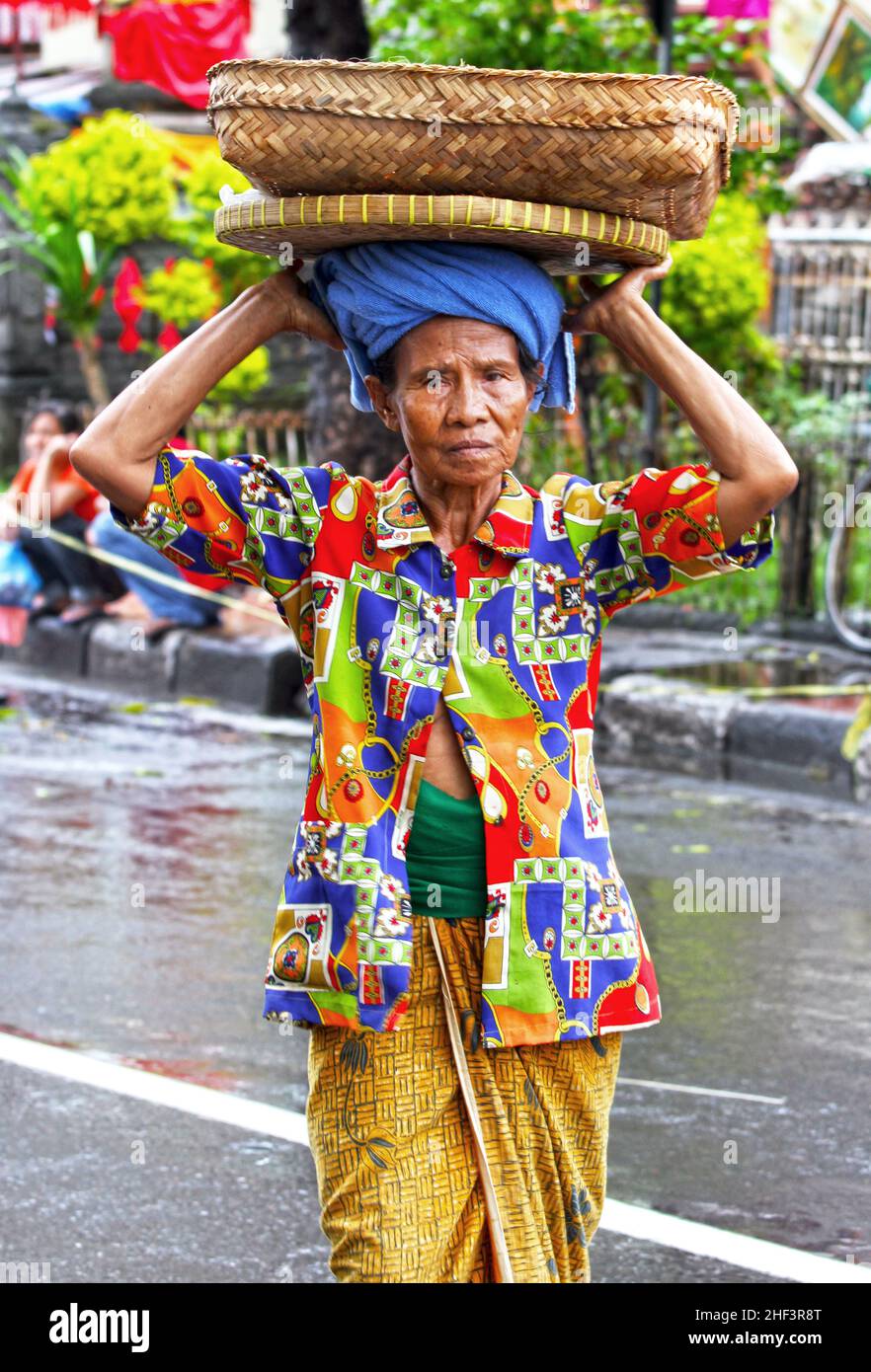 Eine ältere balinesische Frau mit einer bunten Bluse, die einen Korb auf dem Kopf auf dem Sukawati Markt in Bali, Indonesien trägt Stockfoto