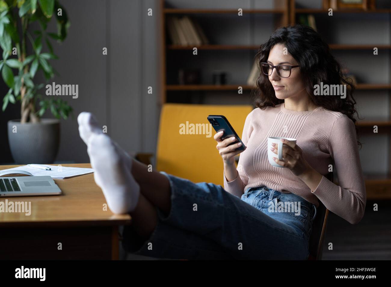 Eine Frau, die an einem entfernten Standort arbeitet, zögerte am Arbeitsplatz, las Text auf dem Mobiltelefon und lenkte sich von der Arbeit am Laptop ab Stockfoto