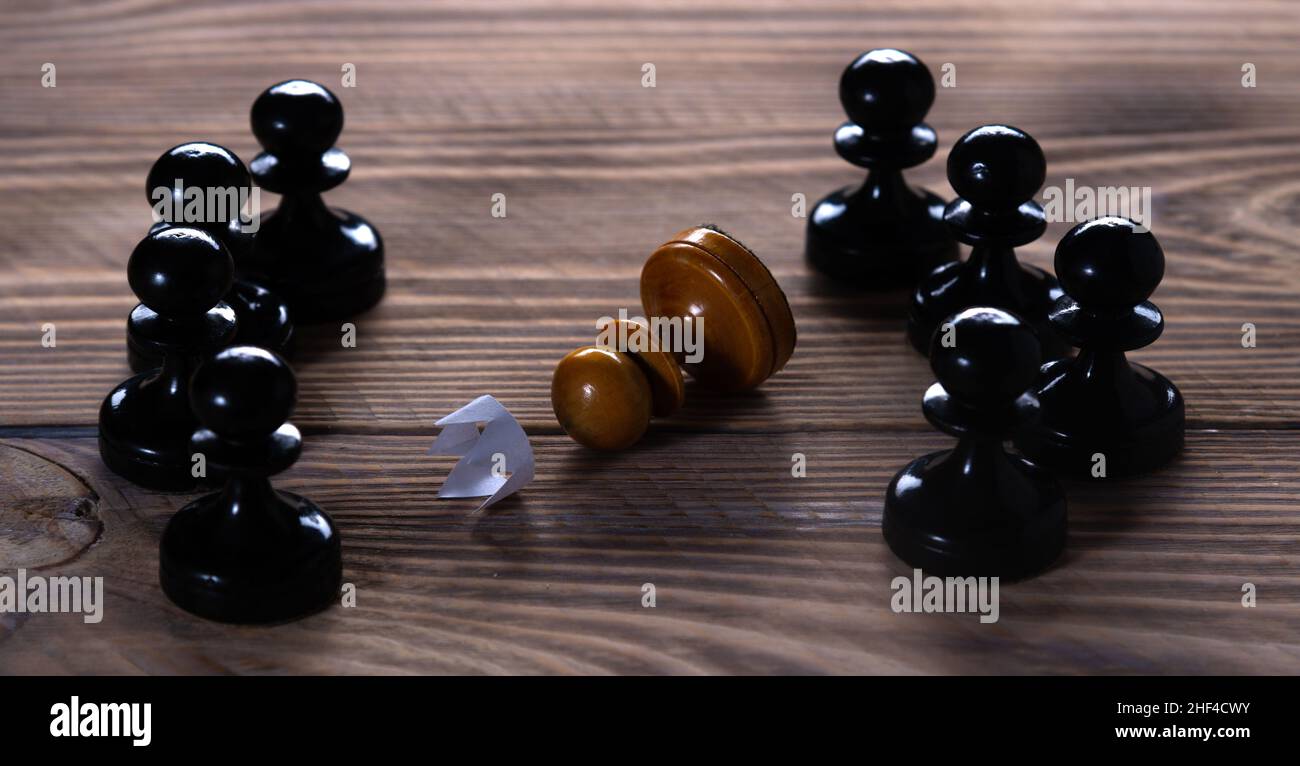 Eine einfarbige weiße Spielfigur mit einer abgelegten Papierkrone liegt umgeben von vielen schwarzen Spielfiguren. Besiegt. Stockfoto
