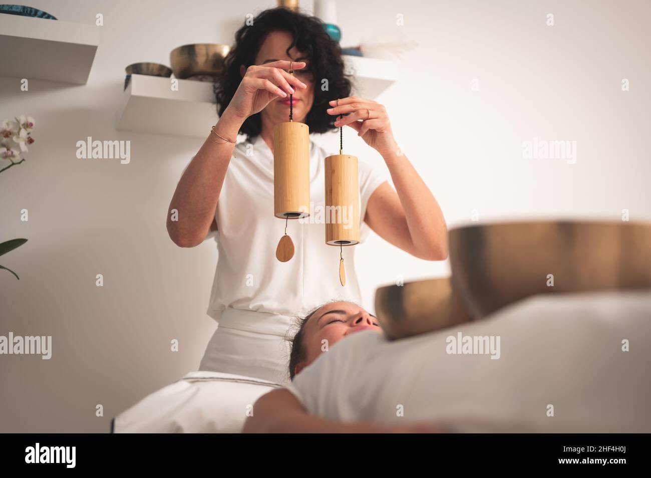 Kaukasische Therapeutin mit Koshi-Glockenspiel-Musikinstrument über dem Kopf einer jungen Frau bei einer Klangheilung Stockfoto