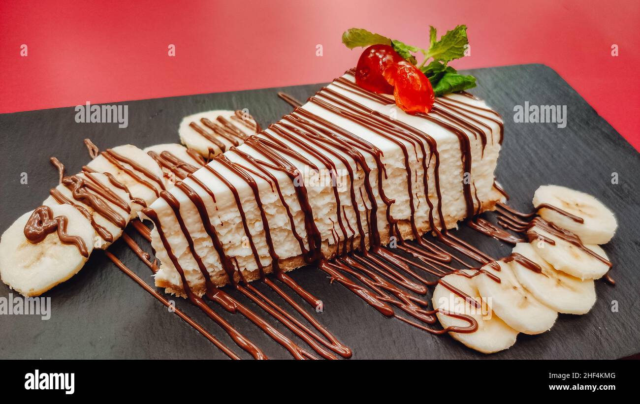 Nahaufnahme eines köstlichen Stückes hausgemachtem Käsekuchen mit Schokolade und Früchten auf Schiefer und rotem Hintergrund. Kochen Dessert und Kuchen Konzept Stockfoto