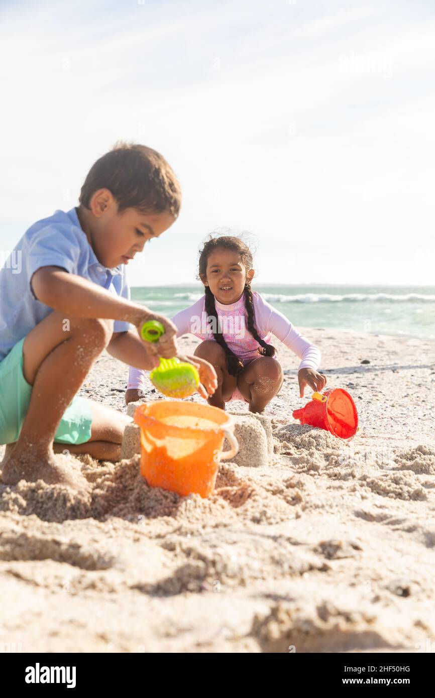 Biracial Bruder und Schwester sammeln Sand in Eimern, während am Strand am sonnigen Tag spielen Stockfoto