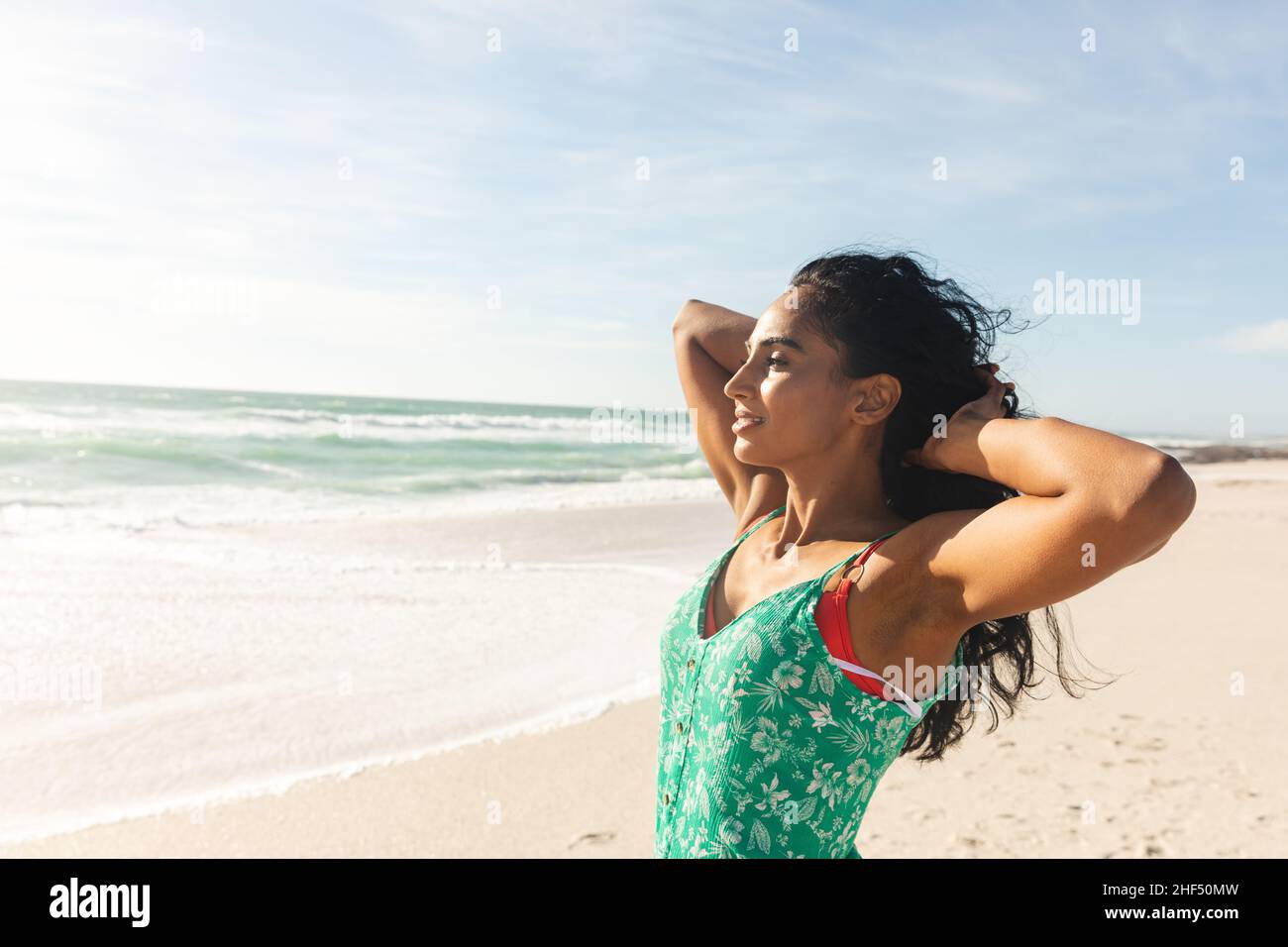 Junge Birazialfrau mit Händen im Haar, die am sonnigen Tag den Strand gegen den Himmel betrachtet Stockfoto