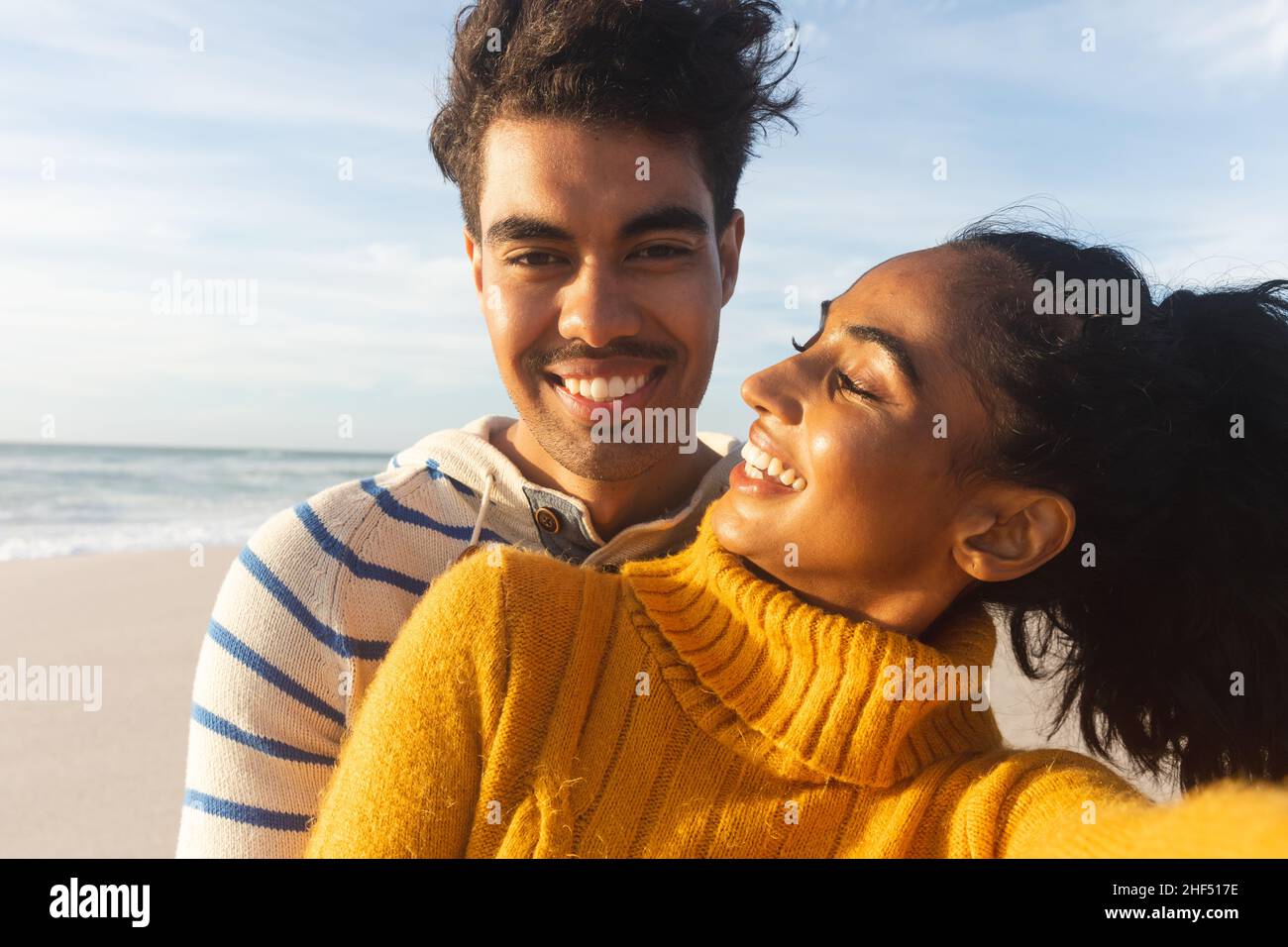 Porträt eines lächelnden Birazialmannes, der am sonnigen Tag eine glückliche junge Freundin von hinten am Strand umarmt Stockfoto