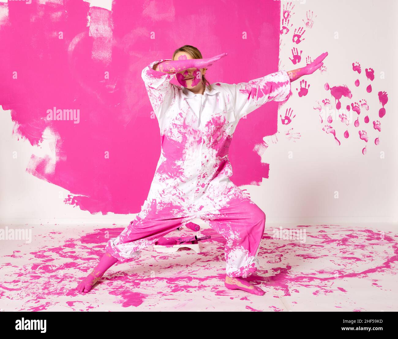 Junges Mädchen, das Spaß hat, eine Wand mit rosa Farbe zu malen. An der Wand hinter ihr sind Spuren von Händen und Füßen zu sehen. Sie trägt Arbeitskleidung und er Stockfoto