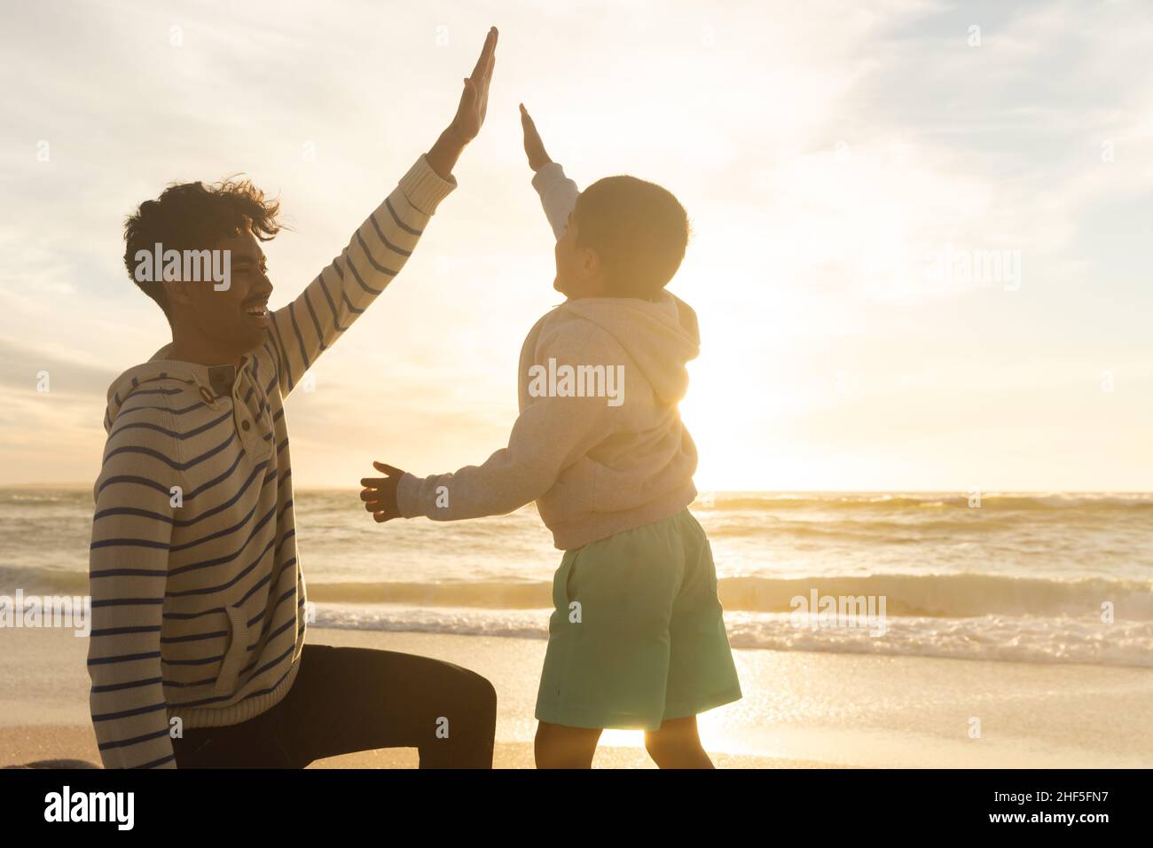 Biracial Junge geben High-five lächelnden Vater kniet am Strand gegen Himmel während des Sonnenuntergangs Stockfoto