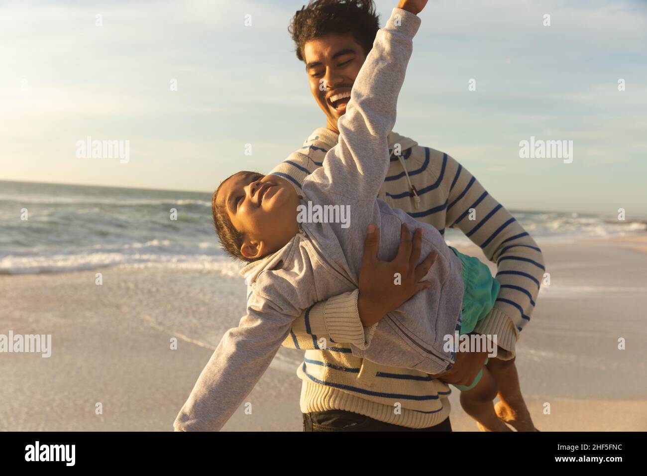 Fröhlicher Birazialmann, der seine Tochter trägt und vorgibt, an sonnigen Tagen am Strand gegen den Himmel zu fliegen Stockfoto Fröhlicher Birazialmann, der seine Tochter trägt und vorgibt, an sonnigen Tagen am Strand gegen den Himmel zu fliegen Stockfoto