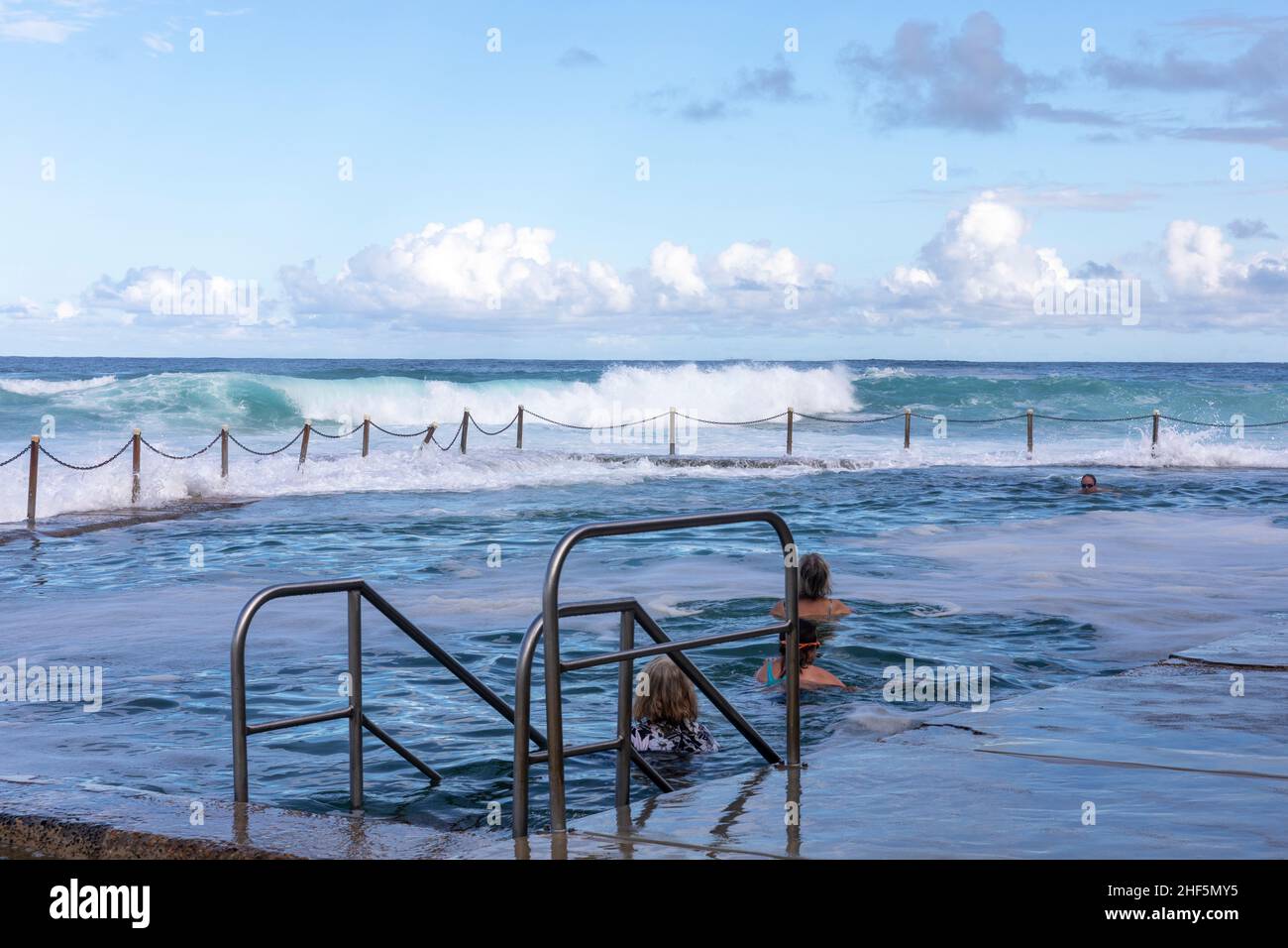 Zwei Damen bereiten sich darauf vor, im Avalon Beach Ocean Rock Pool zu schwimmen, während am späten Nachmittag große Wellen in den Pool und den Strand, Sydney, Australien, stürzen Stockfoto