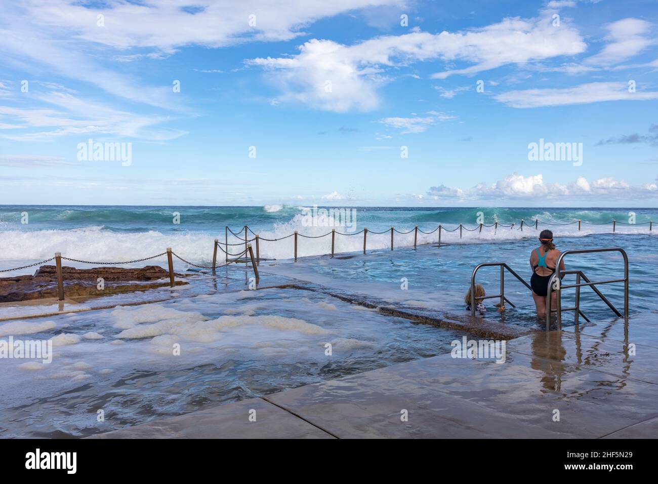 Zwei Damen bereiten sich darauf vor, im Avalon Beach Ocean Rock Pool zu schwimmen, während am späten Nachmittag große Wellen in den Pool und den Strand, Sydney, Australien, stürzen Stockfoto