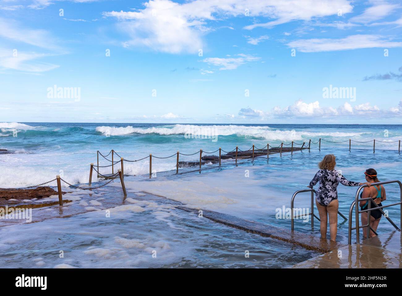 Zwei Damen bereiten sich darauf vor, im Avalon Beach Ocean Rock Pool zu schwimmen, während am späten Nachmittag große Wellen in den Pool und den Strand, Sydney, Australien, stürzen Stockfoto
