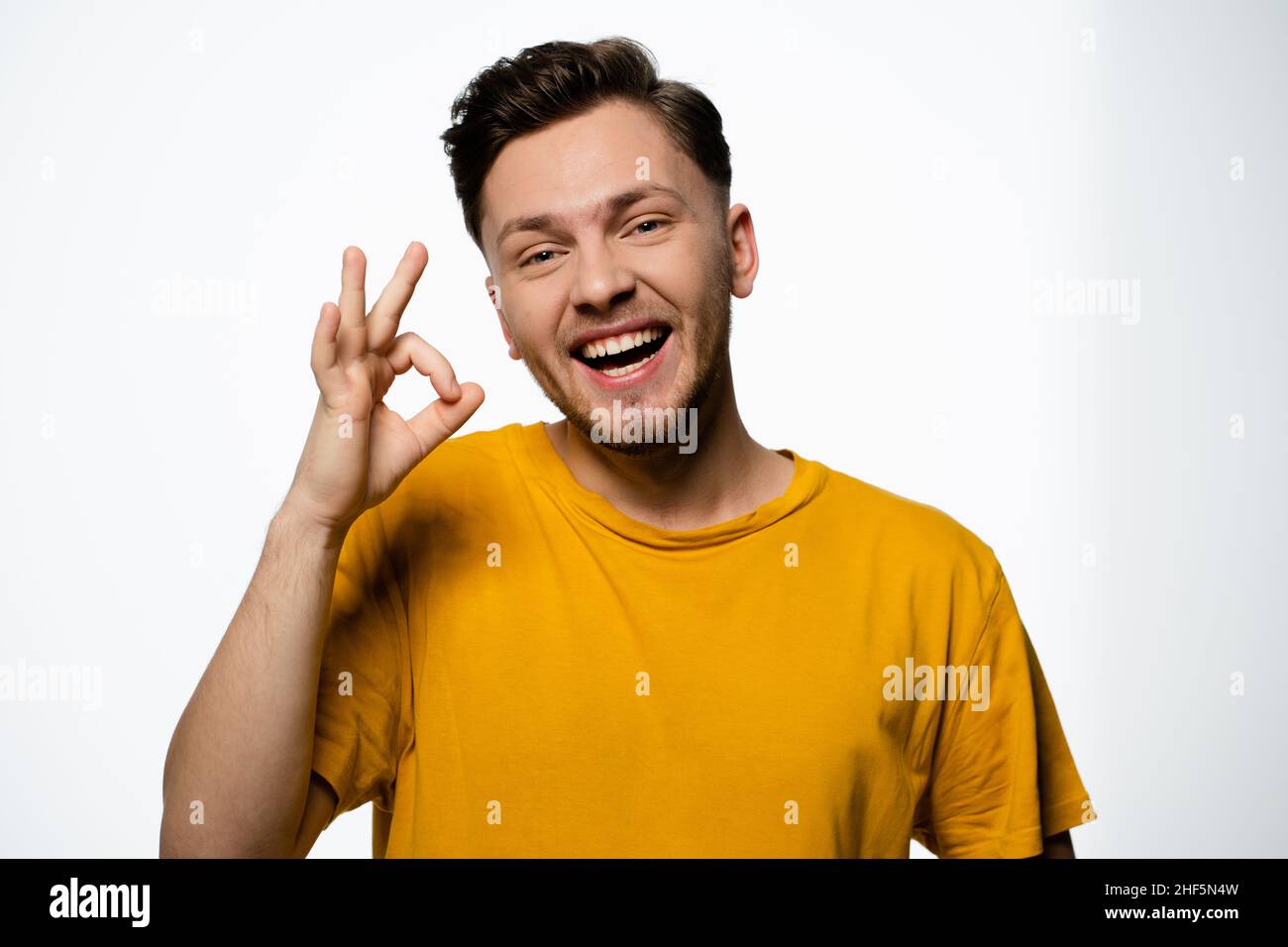 Fröhlicher Mann in einem gelben T-Shirt zeigt eine Geste von Okey auf weißem Hintergrund im Studio. Positive Geste, Ok-Geste. Speicherplatz Kopieren. Nahaufnahme. Hochwertige Fotos Stockfoto