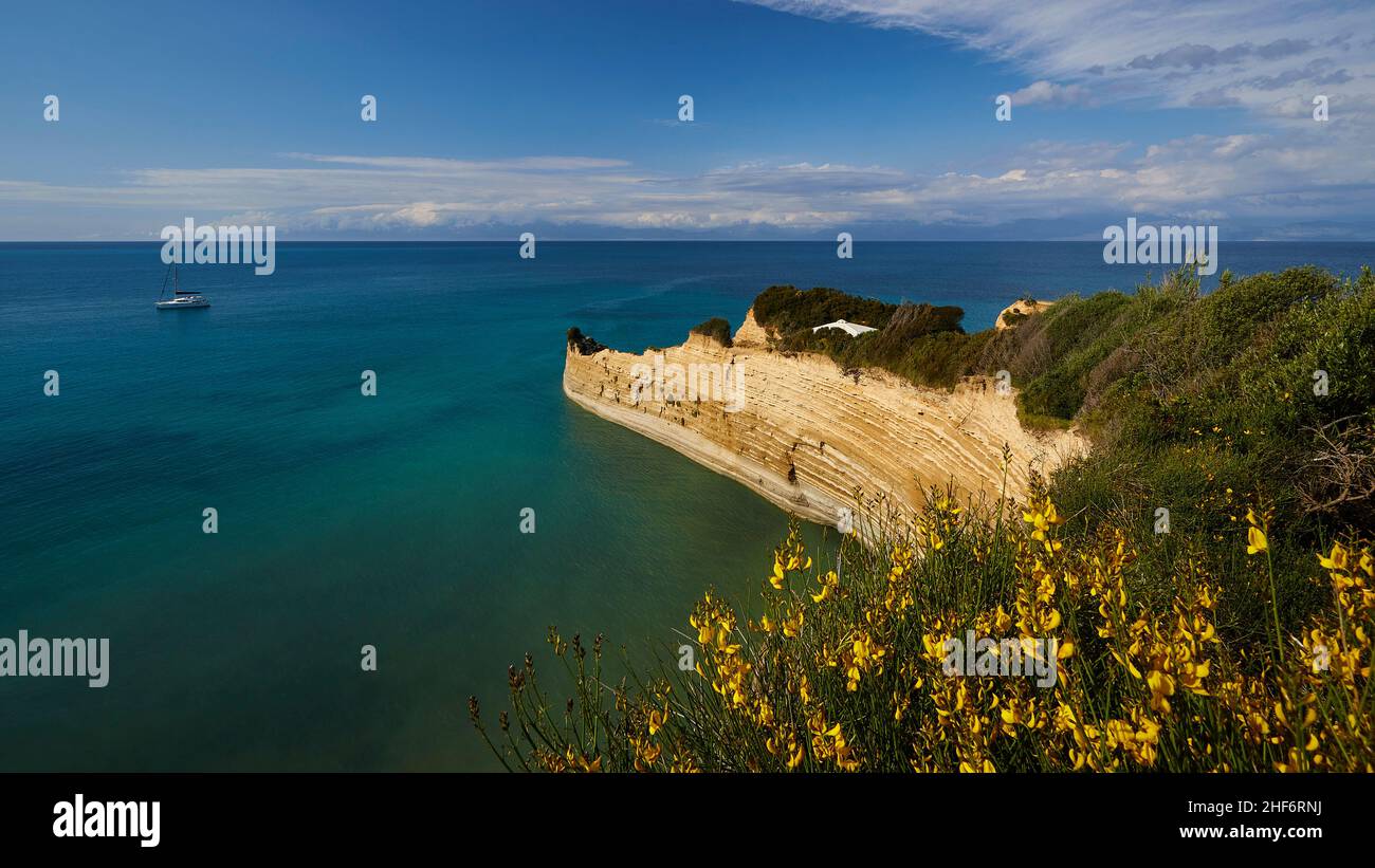 Griechenland, griechische Inseln, Ionische Inseln, Korfu, Nordküste, Bizarre Felsformationen an der Küste, Canal d'amour, blauer Himmel, weiße Wolken, Türkis, Blaues und grünes Meer, Weitwinkelansicht, gelbe Büsche im Vordergrund, ein Segelboot in der Mitte, Blick auf felsige Landzunge Stockfoto