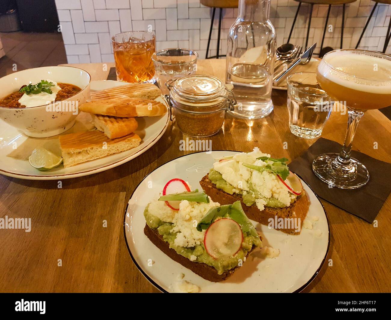Nahaufnahme eines zertrümmerten Avocado-Sandwichs mit Rettich auf Roggenbrot, gegrilltem Brot mit Suppe und zwei Cocktails auf einem Holztisch-Hintergrund. Stockfoto