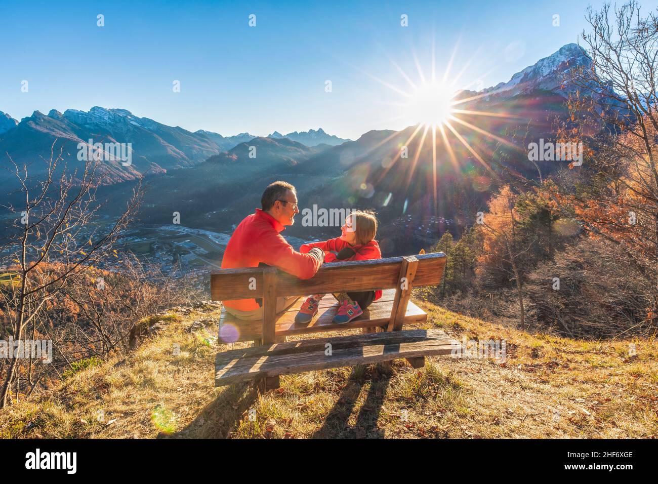Zwei Personen (Vater und Tochter), die auf einer Bank sitzen, lachen und blicken in die Augen, während die Sonne hinter ihnen untergeht, Agordo, Provinz Belluno, Venetien, Italien Stockfoto