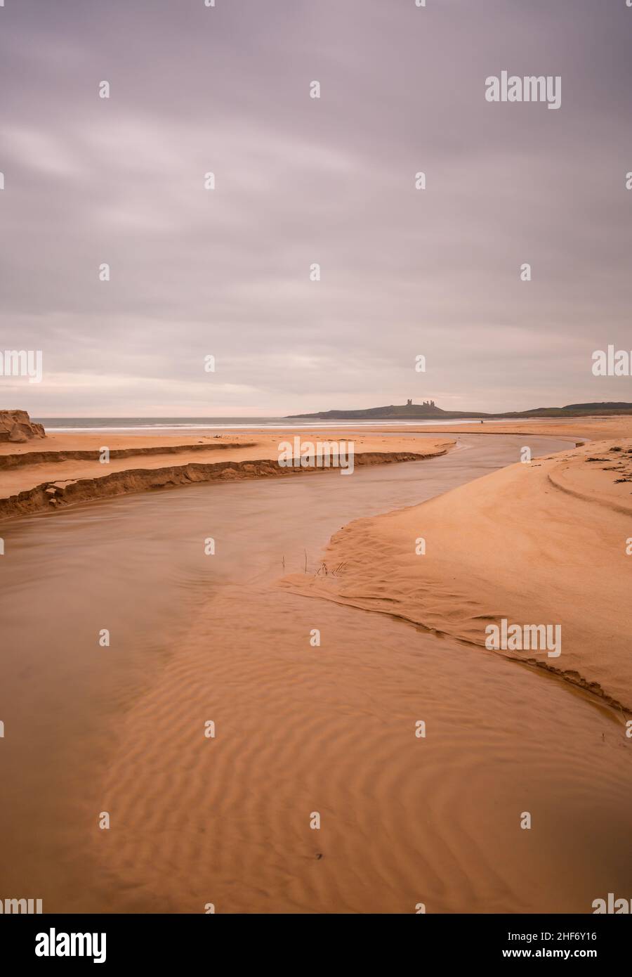 Dunstanburgh Castle in Northumberland, England, aufgenommen mit dem Fluss, der sich im Vordergrund schlängelt und über dem wolkenlosen Himmel liegt Stockfoto