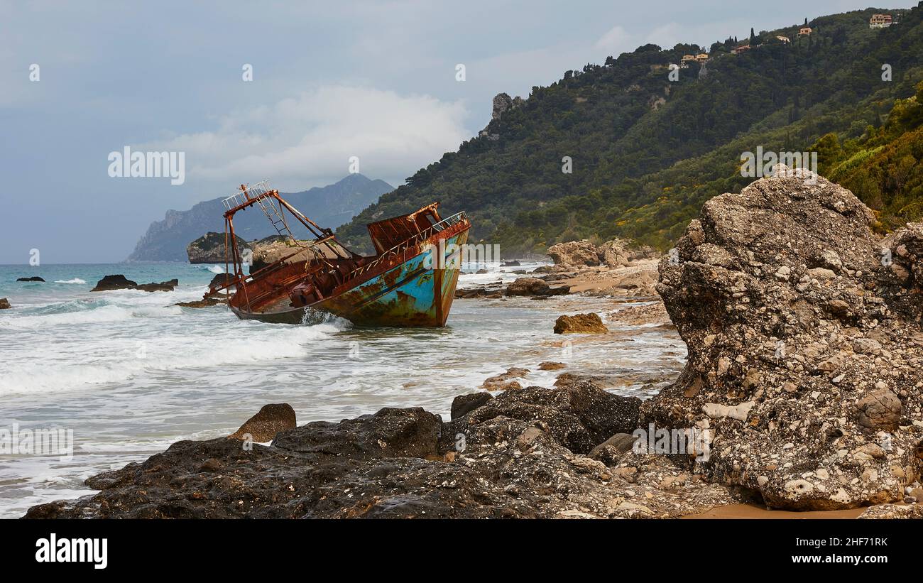 Griechenland, griechische Inseln, Ionische Inseln, Korfu, Westküste, Agios Gordios, schlechtes Wetter, wolkiger Himmel, Schiffswrack, Graffiti-Malerei, Surf, im Vordergrund rechts Felsblock, in der Mitte bunt bemalt, rostig Schiffswrack, dahinter rechts grüne Hügel, im Hintergrund Berge im Norden Stockfoto