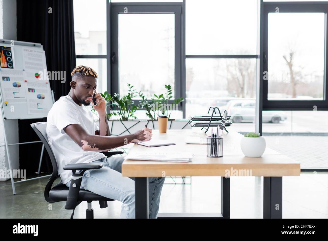 afroamerikanischer Mann mit Vitiligo, der im Büro in der Nähe von Laptop und Flipchart auf dem Mobiltelefon sprach Stockfoto