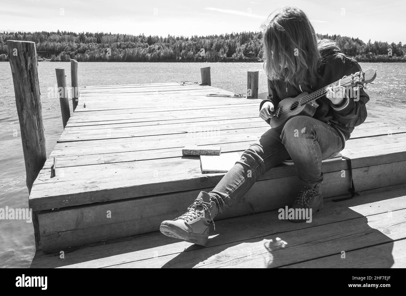 Teenager-Mädchen spielt Ukulele sitzen auf einem hölzernen Pier, schwarz-weiß-Foto im Freien Stockfoto