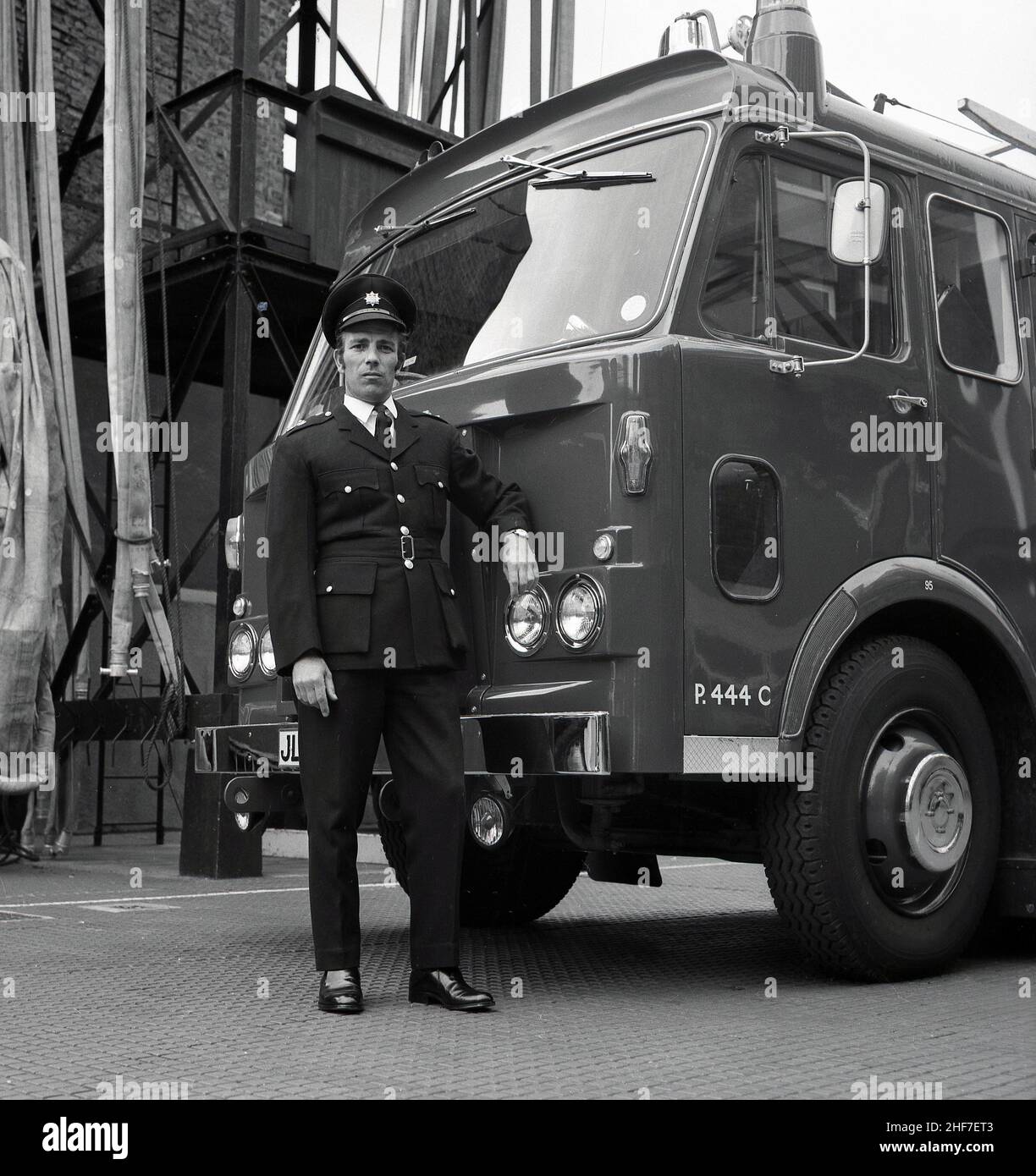 1960s, historisch, ein Stationsbeamter der Londoner Feuerwehr, der in seiner Uniform von einem Dennis-Feuerwehrmann in der Lewisham Fire Station, Southeast London, England, Großbritannien, stand. Die Brüder Dennis, John und Raymond, begannen 1895 mit der Herstellung und dem Verkauf von Fahrrädern, bevor sie zu Autos und Nutzfahrzeugen und dem legendären Feuerwehrauto weiterkamen. Stockfoto